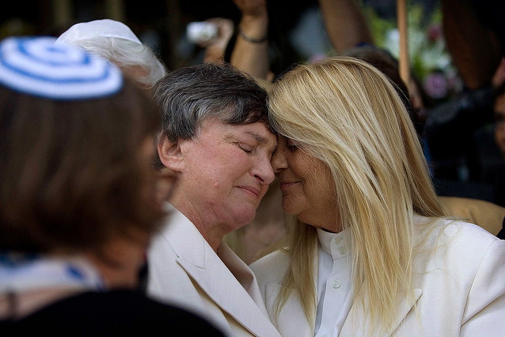 Two women with their eyes closed, embracing. A rabbi's kippah in the background.