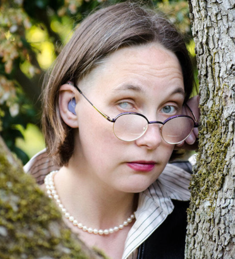 White woman with one clear blue eye and one milky eye, glasses halfway down her nose, a purple hearing aid, pink lipstick, pearls, a button down shirt, leaning against a tree looking like if you push her too hard she's going to duel you and win and it won't be pretty. 