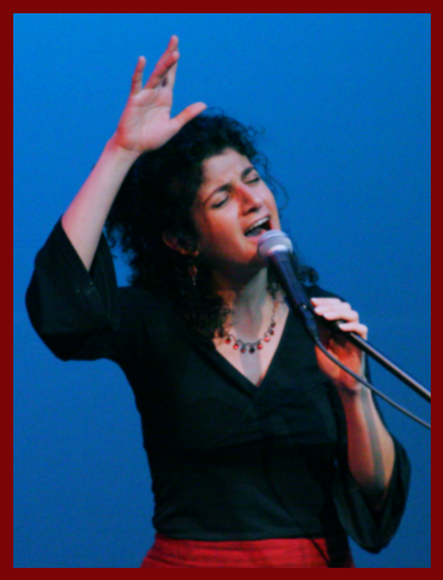 White skinned, curly dark haired woman singing with her eyes closed into the mic, hand raised