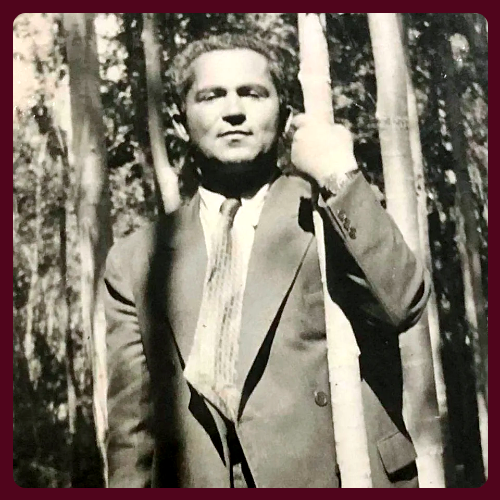 Black and white image of a man in a suit in the trees, half-smiling