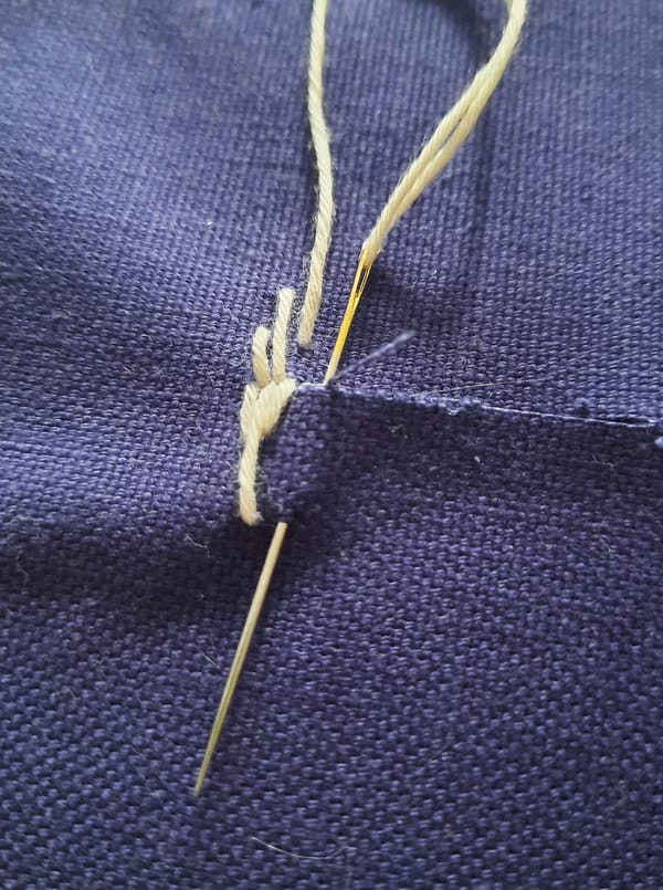 blue fabric with a tear, being mended with white thread and needle