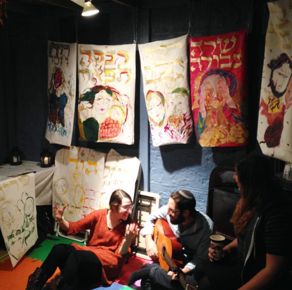 Two people sitting in a sukkah, one with guitar and kippah and beard, amongst gorgeous art depicting biblical figures